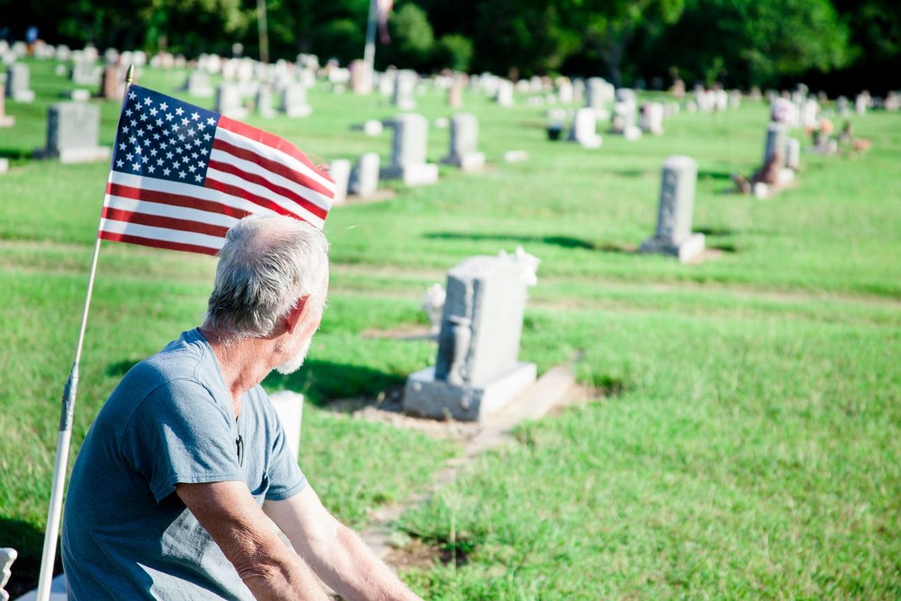 Older adult resting near a cemetery with an American flag